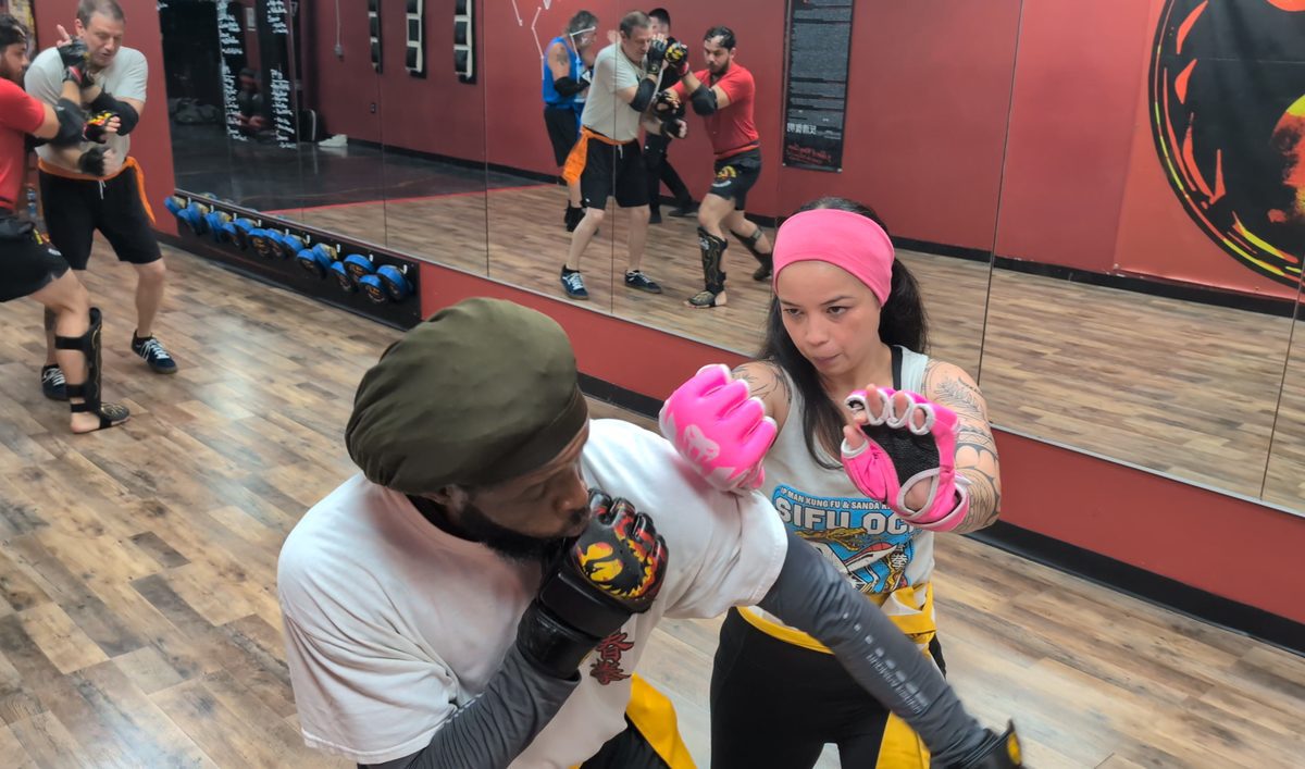 Student in pink gloves practicing a Wing Chun guard drill with a partner while the class trains in the mirror behind them